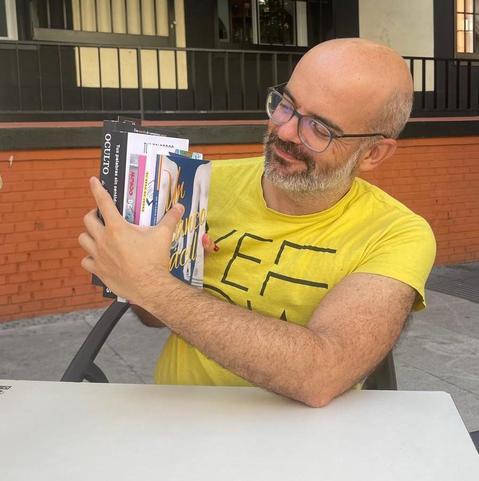 calvo con gafas sentado en mesa de terraza, con camiseta amarilla, enseña orgulloso y sonriente sus libros