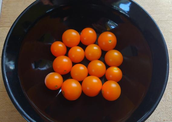 Tiny orange tomatoes in a black ceramic bowl on a cutting board.
