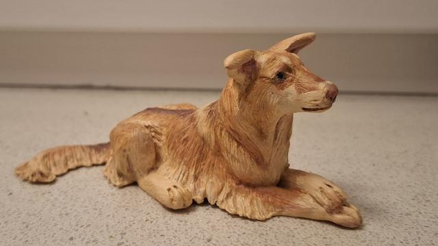 A small clay figurine of a longhaired red liver domino dog with ice blue eyes and slightly floppy ears, laying down with its forelegs crossed daintily.