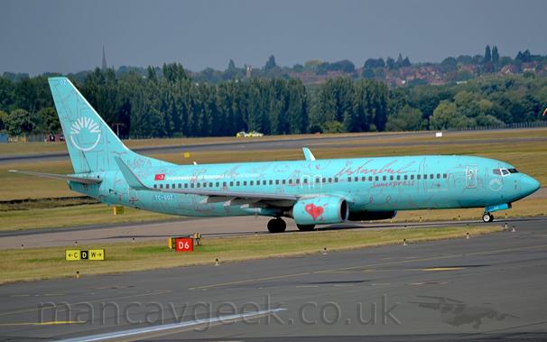 Side view of a twin engined jet airliner taxiing from left to right.
The plane is a very pale blue, with red, seemingly hand-written "Istanbul" titles on the upper forward fuselage, above the forward cabin windows, with additional "SunExpress" below the windows.
Hand-drawn red images of buildings, bridges, vegetation, weather, and curly squiggles cover the rest of the airframe, with a large red love-heart on the side of the engine pods.
There is white text "Impressions of Istanbul by SunExpress" next to a small Turkish flag on the upper rear fuselage.
A white semi-circle topped with 6 white chevrons themselves arranged in a semi-circle, while a smaller version of this logo appears on the nose.
A black taxiway fills the foreground, with grass, more taxiways, and a runway in the background lead up to trees in the distance, with houses beyond that, under grey sky.