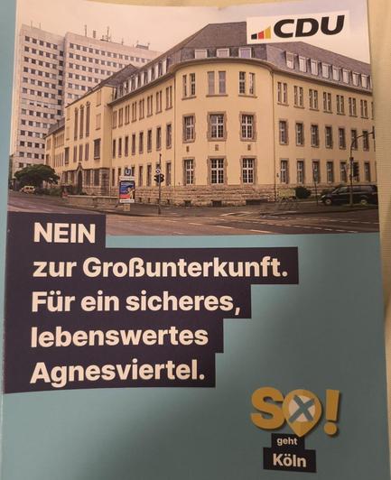 Das Bild zeigt ein Plakat mit dem Logo der CDU (Christlich Demokratische Union) oben rechts. Es zeigt ein Gebäude im Agnesviertel von Köln, mit einem modernen Hochhaus im Hintergrund. Der Text auf dem Plakat lautet: "NEIN zur Großunterkunft. Für ein sicheres, lebenswertes Agnesviertel." Unten rechts ist das Logo von "So! geht Köln" zu sehen. Das Plakat drückt eine politische Botschaft aus, die sich gegen die Errichtung einer Großunterkunft in der Nähe des Agnesviertels richtet und für die Sicherheit und Lebensqualität in diesem Stadtteil eintritt.