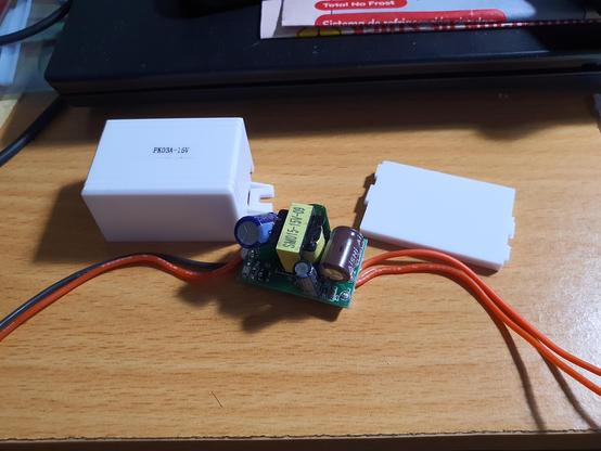 Imaxe dunha pequena fonte de alimentación desmontada e fóra da súa caixa. Aparece o circuito da fonte de alimentación onde destaca un pequeno transformador e dous condensadores electrolíticos ós lados. Xunto coa fonte aparece a caixa de plástico na que vai encerrado o circuito, e a correspondente tapa.