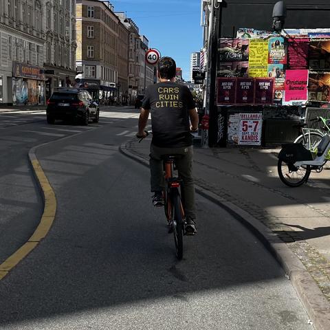 A cyclist wearing a black t-shirt that says "CARS RUN CITIES" rides on a street with a curved yellow line. In the background, there are parked bicycles and various colorful posters on buildings.