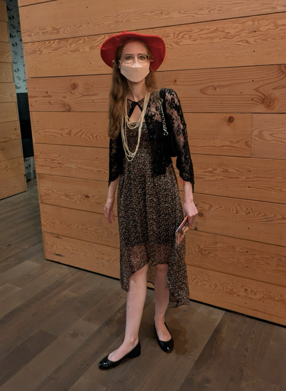 Me posing in front of a wooden wall wearing a fluttery dress with a tiny floral pattern on black. It is short in front and long in back. I have a black beaded garment over it buttoned at the top, a long, pale beaded necklace, black flats, and a large-brimmed red hat.
