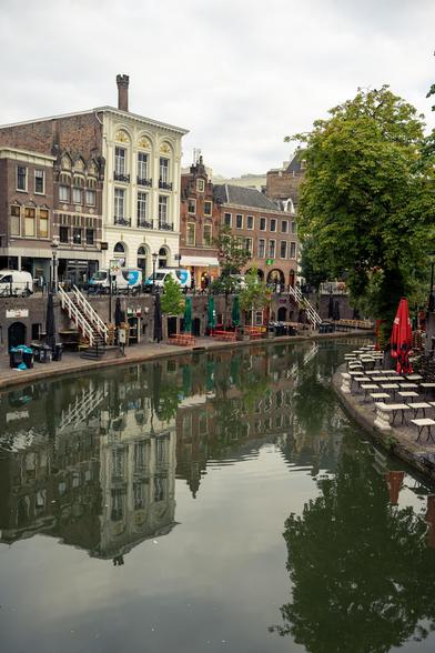 The image shows a peaceful canal scene with buildings lining both sides and several cafes or restaurants visible on the right side. A calm canal runs through the center of the image, flanked by brick buildings with large windows reflecting the overcast sky. On the right side, multiple establishments are present, identifiable by red umbrellas and outdoor seating areas. Green foliage from trees is visible on the right side, adding a natural element to the scene. The water in the canal is still, creating clear reflections of the buildings and trees.