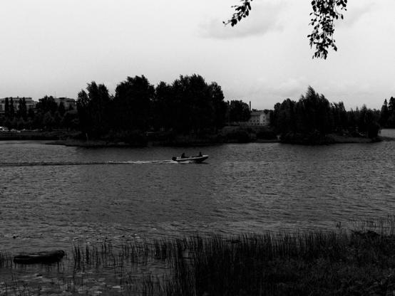 Black-and-white photo of a scene in rainy late-summer day on river. A small boat is speeding across the water. Trees and buildings line the far shore.