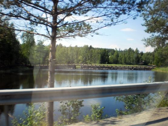 Scenic view through a car window, showing a tranquil waterway surrounded by lush green trees under a clear blue sky. An old bridge lies in the distance. A metal guardrail lines the road.