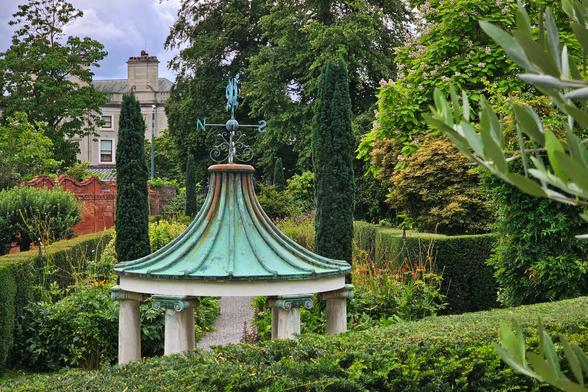 A serene garden scene with a central, circular, dome-shaped copper pavilion featuring a weather vane on top, surrounded by lush green foliage, manicured hedges, and tall cypress trees. In the background, a historic building with a tall chimney is partially visible through the trees. The overall mood is peaceful and tranquil.