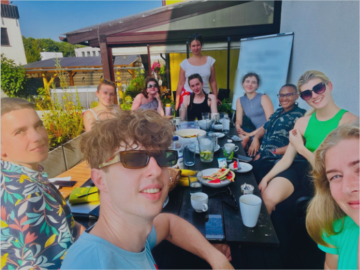 This image shows the whole 10 members of the Inspiration Forum team sitting at a large wooden table in the garden. The table is decked in food, plates, cups and mugs, phones, and various beverages and snacks. Behind the table and team in the right corner is a whiteboard with some of the results of a successful brainstorming session.