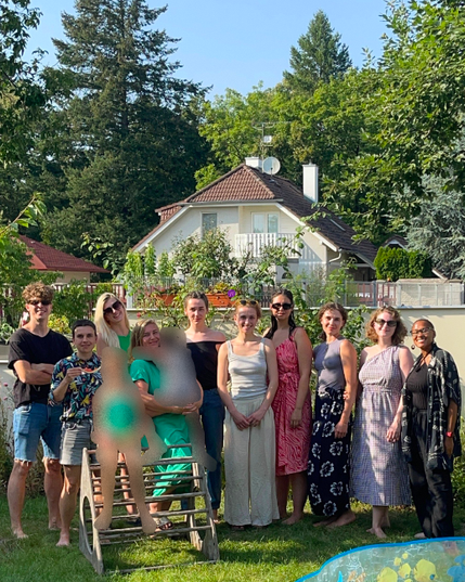 This image shows the entire team of 10 people standing in a garden, smiling a the camera. Two children in the image are intentionally blurred. In the background there are plants, flowers, tall green trees, blue skies, a garden wall, and a neighbouring white house with a brown roof beyond the wall. At the bottom right corner of the image, a piece of an inflatable pool can be seen