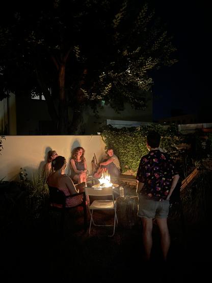 This image shows 4 members of the team sitting around a campfire in the garden at night. 3 people sit on a bench against a white wall, while the fourth sits on a chair with her back to the camera. In front of the bonfire is an empty white, plastic chair. A fifth person in a patterned black shirt and shorts stands to the right with their back to the camera.