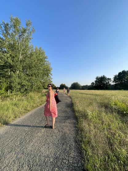 The image was taken as the team is walking back form a day at the lake. Vast blue skies, tall grass, and green trees make up the background of the image. Three members of our team can be seen walking down a concrete path in the distance with their backs turned to the camera. Closer to the camera is Nikol– our Inspiration Forum coordinator– wearing a pink, zebra-print dress and black tote bag, turning back over her left shoulder and smiling into the camera.