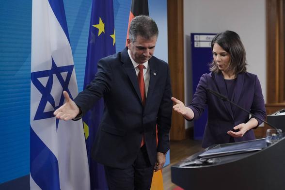 A white man and a white woman in suits. They each offer a palm and look down as a show of grace.
In the background are flags or israel, EU, Germany.

Außenministerin Annalena Baerbock und der israelische Amtskollegen Eli Cohen in Berlin, Feb 2023.