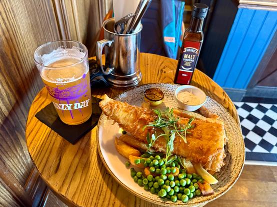 A small table with a pint of beer and a full plate of battered fish, thick fries, peas and a malt vinegar bottle.