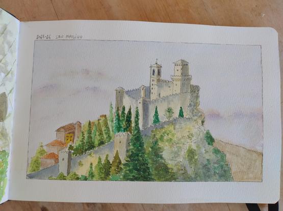 Un acquerello su un quaderno da disegno raffigura la Rocca di San Marino, una fortezza medievale in cima a una collina rocciosa. La rocca, di colore chiaro, ha alte mura merlate e due torri. Ai piedi della fortezza, un bosco svetta verso il cielo, parzialmente coprendo le mura. In basso a sinistra si intravedono alcune case rosse. Il cielo, sullo sfondo, è un velo di nuvole rosa e viola.