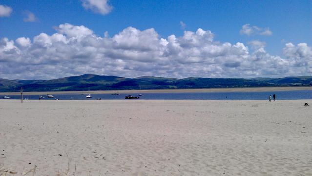 This shows a serene sandy beach stretching towards a calm body of water, with gentle waves meeting the shore. A few small boats float peacefully on the water, their white hulls contrasting against the deep blue. In the distance, rolling green hills rise under a vibrant sky dotted with fluffy white clouds, casting soft shadows over the landscape. The beach appears quiet and tranquil, with only a couple of people walking along the shoreline, adding a sense of scale to the vastness of the scene. The clear blue sky meets the horizon seamlessly, creating a harmonious blend of natural colors—beige sand, blue water, and green hills. A subtle breeze seems to ripple through the scene, hinted at by the faint movement of the distant boats. The overall atmosphere feels calm and inviting, perfect for a peaceful retreat.