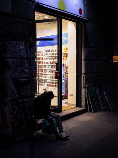 A man sits on a chair outside a shop at night. The shop illuminates the sidewalk, also creating a silhouette of the man, whose details are hidden in the shadows.