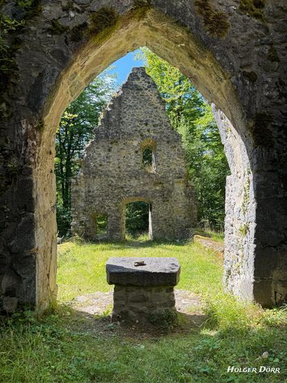 Farbaufnahme aus dem Inneren der Kirchenruine St. Valentin bei Laubach. Der Blick führt vom Bereich hinter dem Altar durch das spitz zulaufende Portal auf eine sonnenbeschienene Wiese und eine gegenüberliegende Ruinenwand mit Fensteröffnung. Moosbedeckte Steine, warmes Licht und sanfte Schatten verleihen der Szene eine ruhige, fast spirituelle Atmosphäre.
