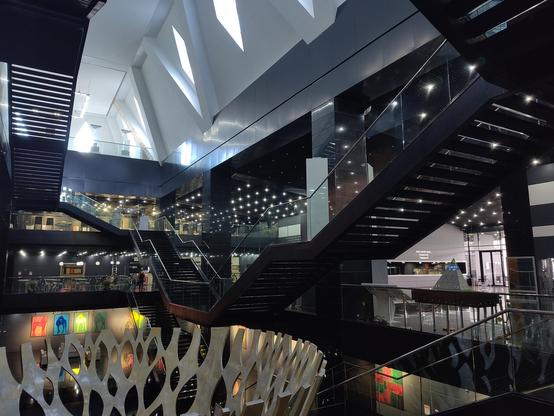 Inside of a pyramid shaped building. Crazy angles, colours and reflections. Stainless steel tree sculpture, black stairways, shiny polished black floors, bright white lights reflected in floors and on walls. Bright coloured paintings. Above, white walls with triangular windows, peace dove images therein.
