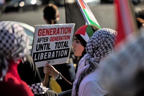 A woman on a street corner looking out towards the road, wearing a serious expression on her face, surrounded by a crowd with a Palestine flag around her head, and holding a sign that says, "generation after generation until total liberation".