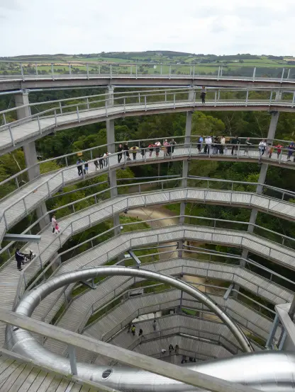 Looking down into the middle of a large spiral structure. Lines if people are walking up the spiral ramp. A stainless steel slide goes down the middle of the structure. Views of forest in the background.