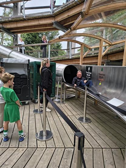 A man gets to the bottom of a stainless steel slide. He is watched by a couple of children (not his).