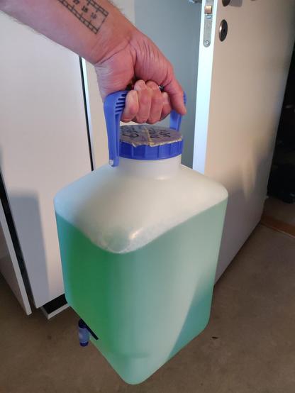 A photo of a hand holding up a 10 litre can with a spout on the bottom left. It is almost full with a green liquid.