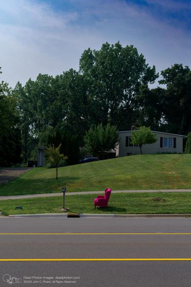 A photograph of a magenta armchair sitting on a grassy verge next to a road with painted yellow lines. A house is on a hill in the background, with large trees and a partly-cloudy sky.