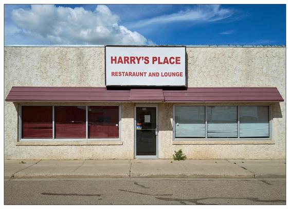 Exterior view of "Harry's Place Restaraunt and Lounge," featuring a single-story building with weathered white stucco and a flat roof. A large red and white sign with the restaurant's name is mounted on the facade above red and maroon awnings shading the windows. The building's left side has a red-paneled window while the right has a larger window with horizontal blinds. A centered glass door provides entrance, under a clear sky with light cloud cover.