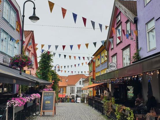 The picture shows a small street. On the left and right are colourful house facades, with flowers and seating areas in front of them. Between the houses there are multi-coloured pennant garlands. The sky is light grey.