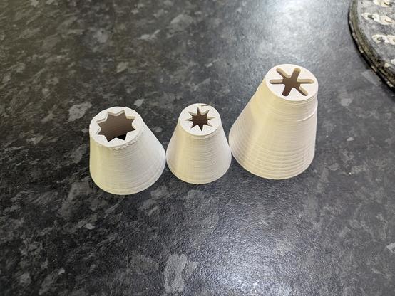 A photo of three churro nozzles on a kitchen bench. There are a variety of shapes at the end of the nozzles with differing numbers of points on the stars and diameters of opening. The nozzles are made of white ABS plastic.