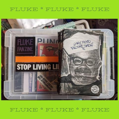 Several copies of "Gary Floyd — picture show" rest on top of a plastic box full of zines. The zine cover shows Gary, a smiling white man with glasses and a beard. He's wearing a sailor hat.
