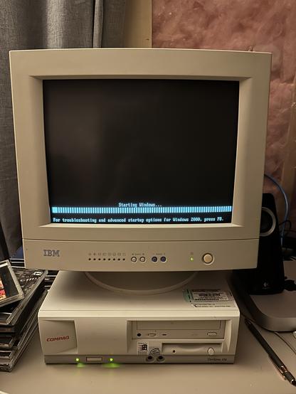 A beige IBM CRT monitor sits atop a Compaq Deskpro EN desktop computer, both part of a retro computing setup. The screen displays the early boot screen of Windows 2000 Professional, showing the “Starting Windows…” text with a horizontal progress bar made of small blue rectangles. Below it reads: “For troubleshooting and advanced startup options for Windows 2000, press F8.” To the left are a few jewel cases for CDs, and to the right is a Logitech speaker and a Mac mini is just barely visible. The background features pink fiberglass insulation and a curtain.