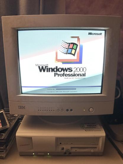 The same IBM CRT monitor and Compaq Deskpro EN are now shown further along in the boot process. The screen displays the Windows 2000 Professional splash screen, featuring the Windows flag logo and the text “Built on NT Technology.” A loading bar near the bottom reads “Starting up…” with a copyright notice below: “Copyright © 1985–1999 Microsoft Corporation.” The glow from the CRT adds a soft light to the otherwise dim workspace. The rest of the setup remains unchanged, including the CD cases, speakers, and nearby Mac mini.