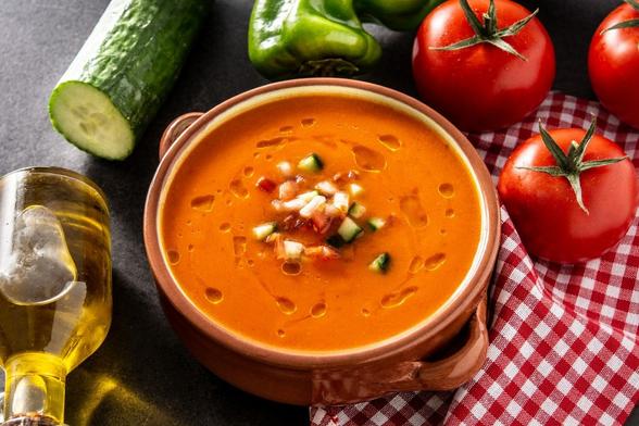 Una cazuela de barro con gazpacho rodeada de alguno de los ingredientes que se usan para hacerlo, como aceite de oliva, pepino, pimiento y tomates.