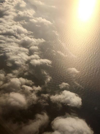 Blick aus einem Flugzeugfenster auf eine wolkenbedeckte Landschaft über dem Meer. Dichter, weißer Wolkenteppich zieht sich schräg von links unten nach oben, während rechts die glitzernde Wasseroberfläche des Ozeans zu sehen ist. Die Sonne reflektiert intensiv auf dem Wasser und erzeugt einen goldenen Schimmer, der das Bild in warmes Licht taucht. Die Wolken wirken bauschig und plastisch, als würden sie knapp über der Meeresoberfläche schweben.