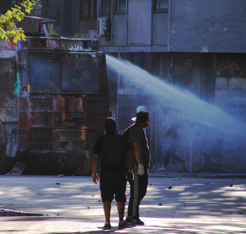 A daytime urban scene showing an armored water cannon (guanaco) covered in graffiti on the left side, firing a powerful, misty white jet of water toward the right. In the foreground, with their backs to the viewer, two men observe the scene; one of them is wearing white gloves and has what appears to be a gas mask hanging around his neck. The street floor is covered in debris and rocks. In the background, urban buildings with more graffiti and some blurred silhouettes of people can be seen amidst the mist.
 This was in the context of the social protests in Chile in 2019.