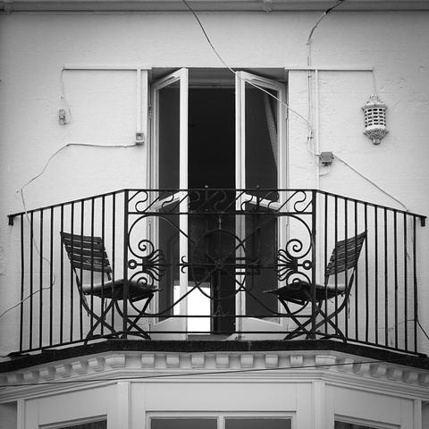 Black and white square image. Two wooden folding chairs with seat cushions, placed at a 90° angle from each other and a 45° angle from the viewer. They are on an otherwise empty upstairs balcony with an open French window style door behind them. Several lengths of wire extend across the wall from the left, the right, and above. An ornate painted metal lamp is near the top right of the picture.