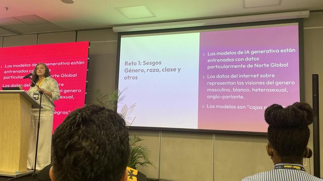 sesgos en los modelos generativos de IA, que aparecen resaltados en una pantalla detrás de él. Los puntos clave incluyen la influencia de los datos de entrenamiento, problemas de representación y críticas a los modelos existentes. Algunos miembros del público son visibles