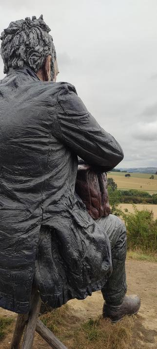 a large sculpture of a man sitting on a three legged stool looking out over open countryside. he has a bag on his knees.