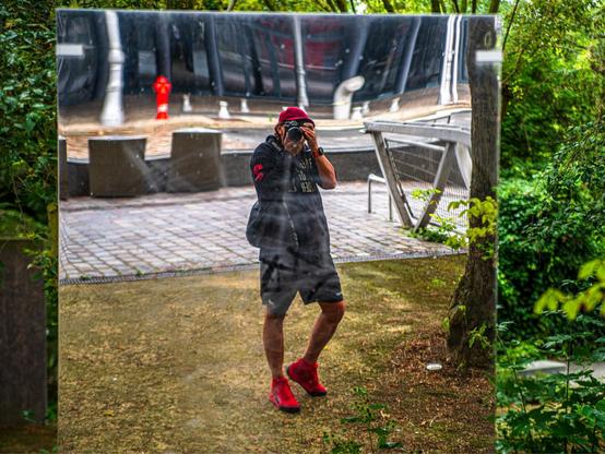 An individual in casual attire is reflected in a large mirror. They are holding a camera, capturing the scene. The background features greenery and a paved area with furniture, along with a fire hydrant visible in the reflection.