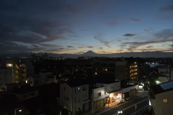 Landscape photograph of a Japanese cityscape just before nightfall, with the singular silhouette of Mount Fuji standing out against the reddish sky at the horizon. The horizon, forming a line at the vertical middle of the picture, is marked by dark cloud shapes to the left and right of the central Mount Fuji.
Above the horizon, the sky fades into a dark blue. Thin dark gray clouds appear to be sprayed on its sides. Under the horizon, the dark residential maze is lighted by street lamps.