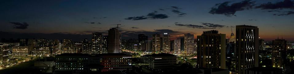 The image captures a cityscape at twilight, showcasing the transition from day to night. Skyscrapers dominate the skyline, their windows illuminated by artificial lighting, contrasting with the darkening sky above. The structures vary in height and design, indicating a mix of commercial and possibly residential buildings. Some skyscrapers are under construction or renovation, as suggested by cranes visible on top. In contrast to these towering edifices, lower-rise buildings can be seen at ground level, providing a sense of scale and urban diversity.

The lighting conditions in the photograph suggest that it was taken during dusk, with the sun having recently set below the horizon but its light still lingering enough to cast some residual illumination on parts of the cityscape. The ambient light is softening as night approaches, contributing to the calm yet dynamic atmosphere of an active metropolis winding down for the evening.