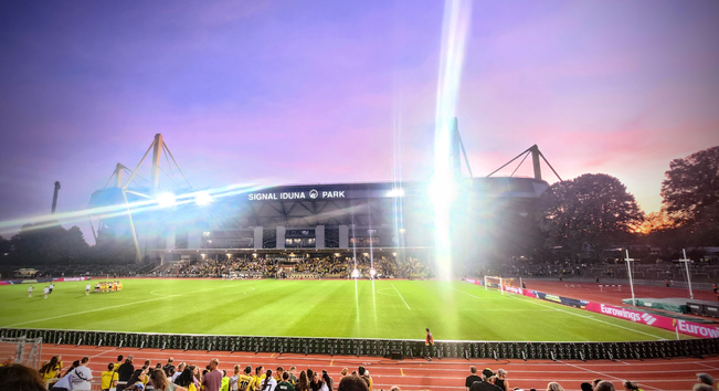 Das Stadion Rote Erde. Das Flutlicht blendet vor dem Abendhimmel. In Hintergrund das Westfalenstadion. Rechts daneben Bäume hinter denen die Sonne am Horizont verschwindet. Links auf dem Feld feiert die siegreiche Mannschaft... eh... Frauschaft...