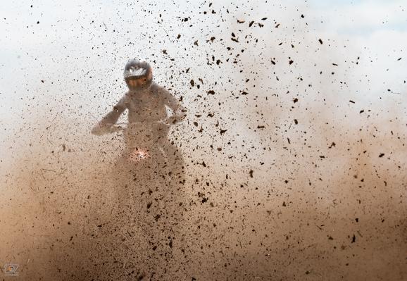Landscape format photo. On the left side of the image, a motocross rider on a motorcycle can be seen dimly behind a cloud of dust and dirt.