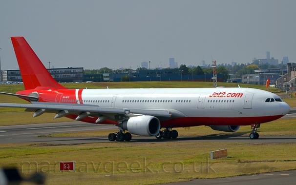 Side view of a twin engined jet airliner taxiing from left to right along a grey taxiway lined by grass.
The plane is largely white, with red belly, rear fuselage, and tail.
There are red "Jet2.com" titles on the upper forward fuselage.
Green grass fills the foreground, with a grassy mound dominating the background with buildings behind it.
A distant city-scape slowly vanishes into hazy grey sky.
