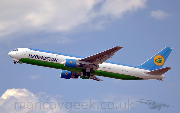 Side view of a twin engined jet freighter howling into the air, flying from right to left with the undercarriage being stowed, flaps extended from the rear of the wings, and the nose raised significantly, suggesting it has just taken off.
The plane has a wide white stripe running along the middle of the body, with blue on the upper body and tail, and green on the belly, both outlined in red.
There are black "Uzbekistan" titles on the lower forward fuselage, and the registration "UK67002" on the lower rear, with additional black text "Boeing 767-300ER" just aft of the wing root.
On the tail, a stylised green flying bird is in the middle of a yellow circle that is outlined in red.
Pale blue sky fills most of the rest of the frame, with the plane threading it's way between fluffs of white cloud at top and bottom.