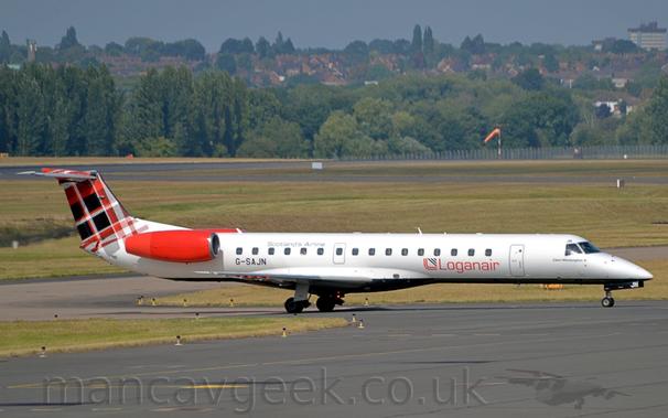 Side view of a twin engined jet airliner taxiing from left to right  along a black taxiway.
The plane is mostly white, with a black belly.
There are red "LoganAir" titles on the lower forward fuselage, with the black registration "G-SAJN" on the lower rear, just in front of and below the red engine pods mounted on the sides of the rear fuselage.
The tail has a red, black, and white tartan pattern.
Black taxiway fills the foreground, lined by green grass on the left.
More grass, black taxiways, and a runway fill most of the background, leading up to trees in the distance, with houses nestled in the trees.
Grey-blue sky fills the rest of the frame.