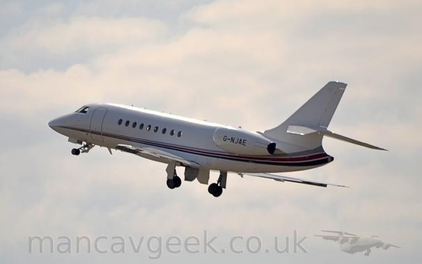 Side view of a twin engined BizJet flying from right to left at a low altitude, with the undercarriage in the process of being stowed away, flaps extended slightly from the rear of the wings, and the nose raised sharply, suggesting it has just taken off.
The plane is mostly white, with  a grey, mauve, and blue stripe running along the body from the nose, getting wider as it moves rearwards and sweeping up into the lower reaches of the tail .
The black registration "G-NJAE" is on the side of the engine pods, mounted on the sides of the rear fuselage.
Strong sunlight is reflecting off the top of the fuselage
Cloudy white sky with patches of blue fill the rest of the frame.