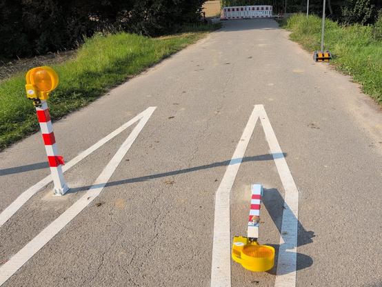 Feldweg. Im Vordergrund zwei Poller mit aufgesetzten Baustellenleuchten, einer davon liegend. Im Hintergrund Baustellenabsperrungen.